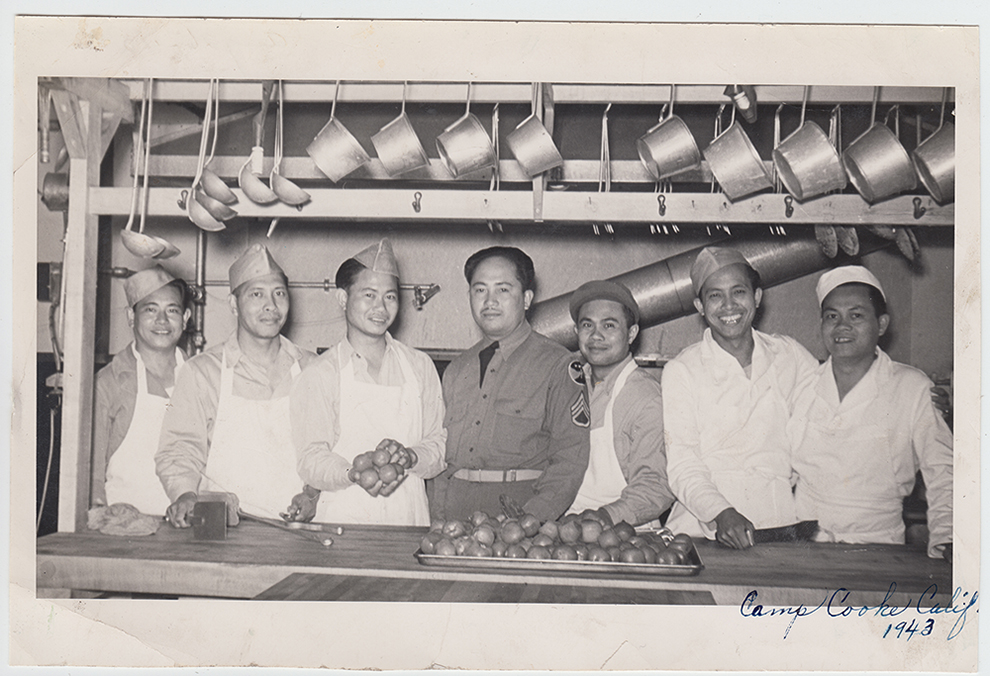 A row of men behind a table in a kitchen, pans hanging above them